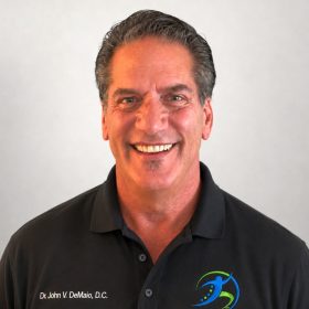 Portrait of Dr. John V. DeMaio, D.C., smiling, wearing a black polo with a blue-green clinic logo on the chest.
